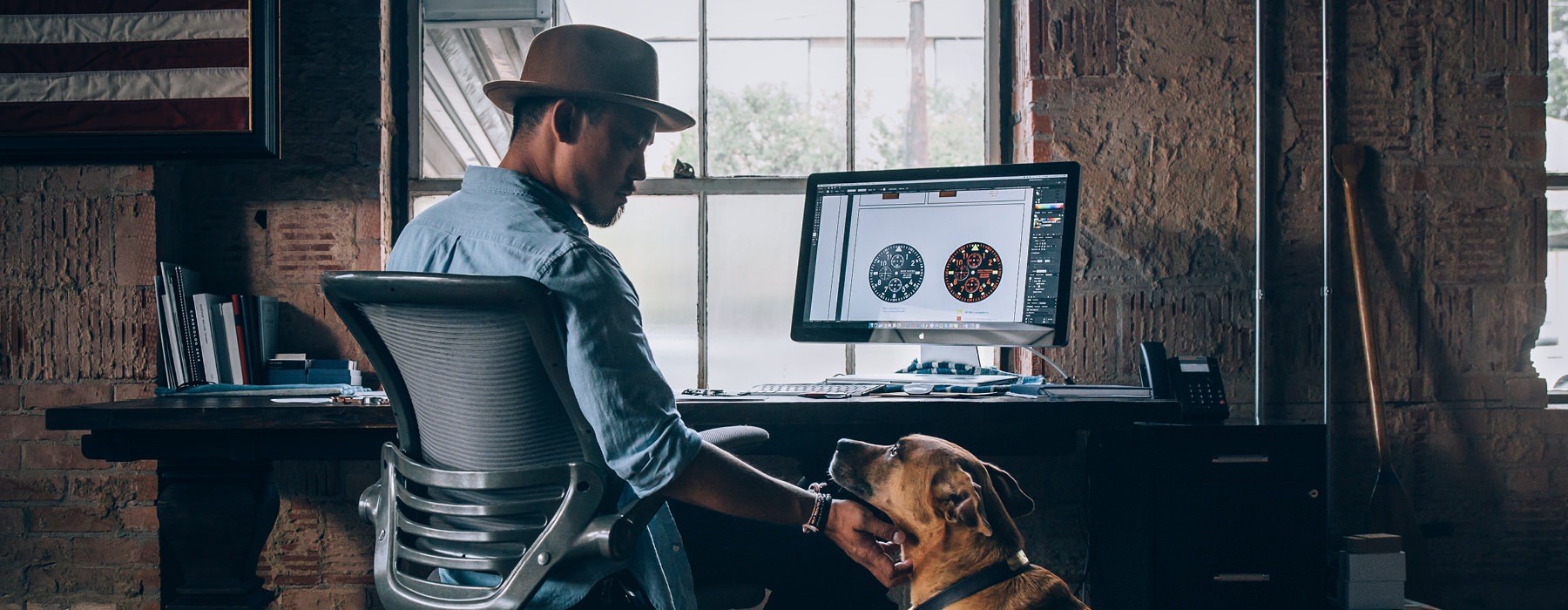 young man with at hat, sits at his desk and scratches his dog's neck as they look at each other
